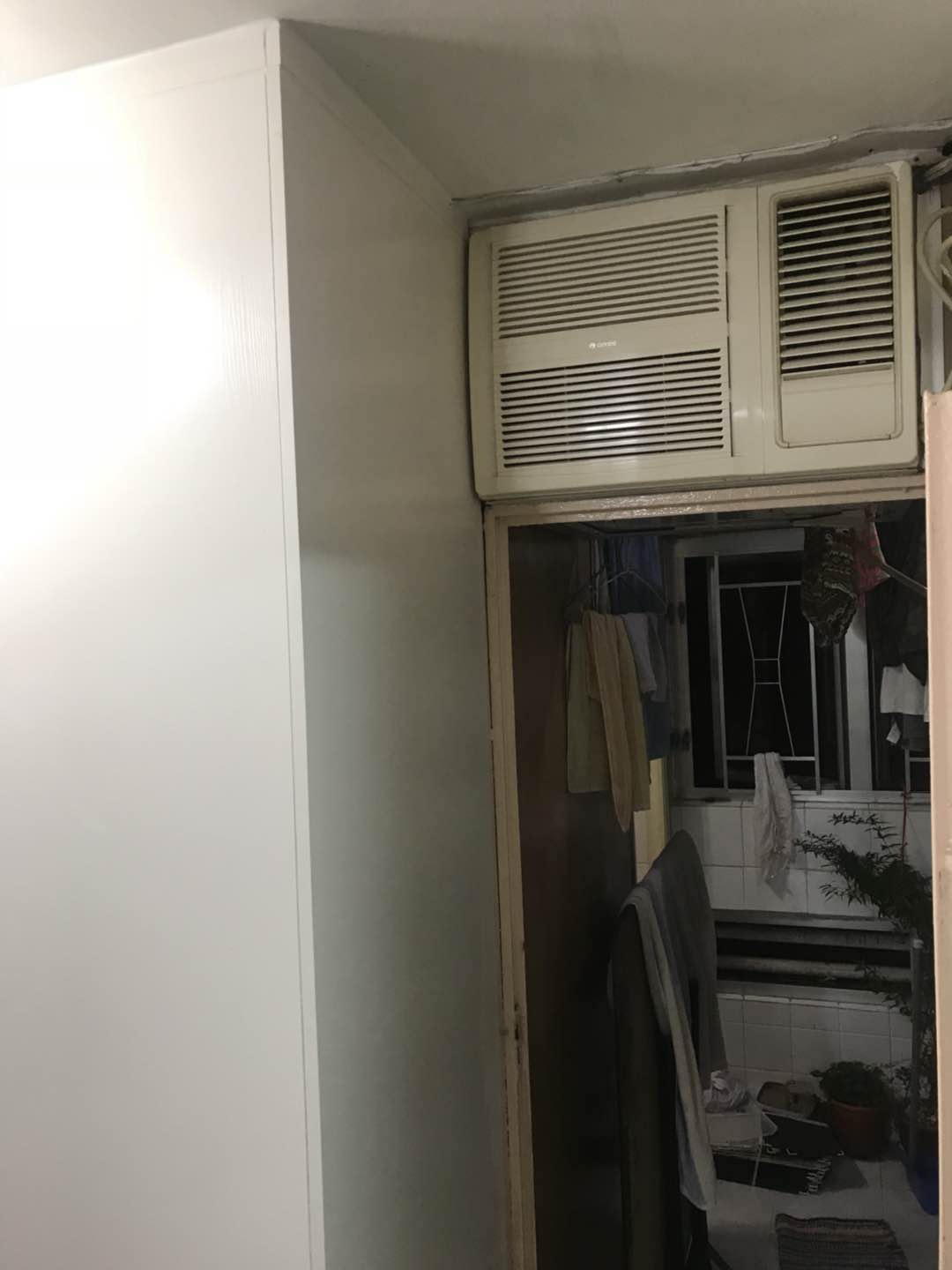 睡房角望向露台,左邊一幅白色到頂訂造雙面櫃側面貼住天花同牆角,右上裝咗白色窗口式冷氣機,下方門口通往晾住淺色毛巾的露台,可見窗框、白瓷磚牆同盆栽,環境光線偏暗。