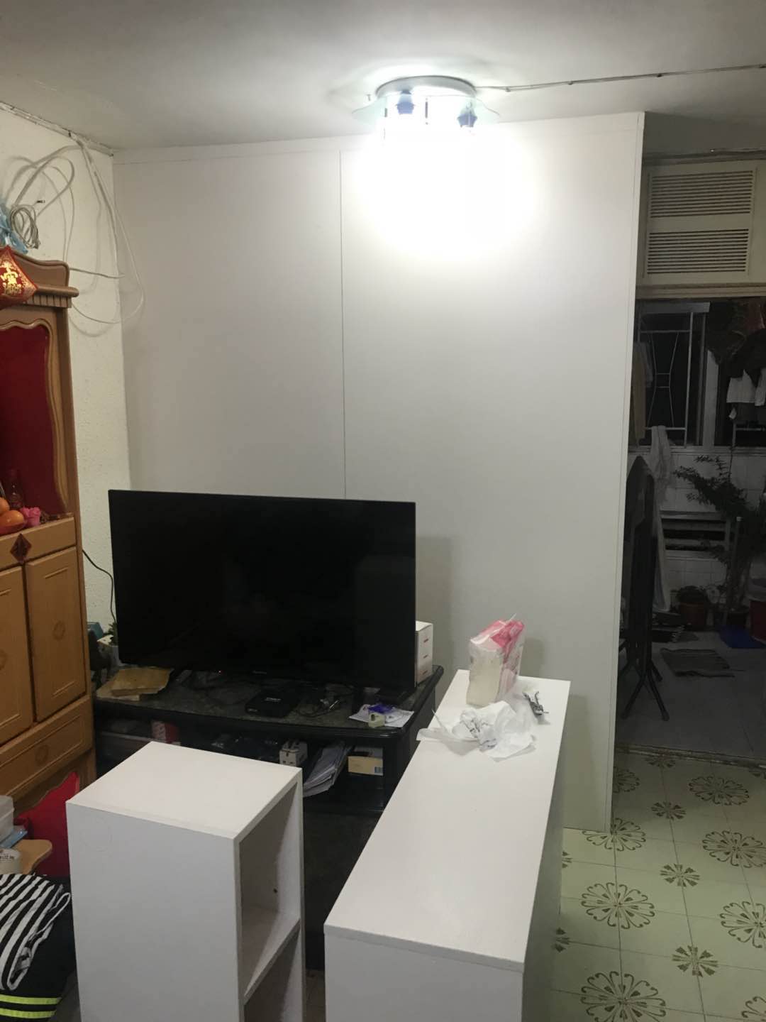 公屋客廳實景,中央一幅白色到頂訂造雙面櫃將空間一分為二,櫃前放咗黑色電視櫃同一部大尺寸電視,旁邊有白色矮櫃,左邊係深木色舊式組合櫃連神位,右邊櫃側留通道望入鋪綠色花磚的露台,天花裝咗圓形射燈。