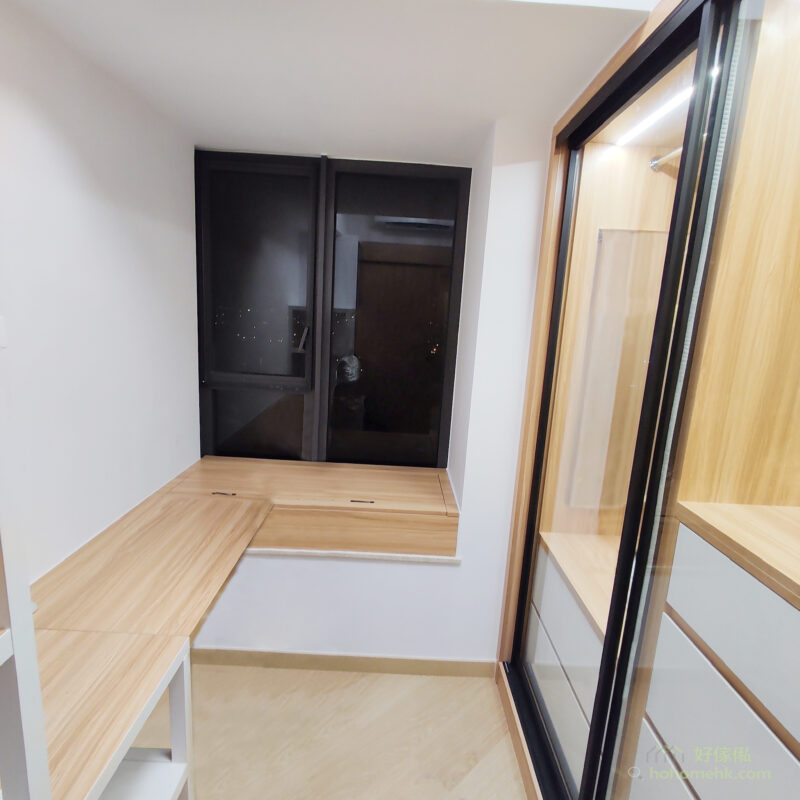 衣帽間窗台一角，左下方係L形淺木色地台座位連收納，靠牆係一組黑色玻璃趟門衣櫃，門上有掛鈎；右邊係高身深框玻璃門衣櫃，櫃內亮起燈條，下方配白色抽屜，地面鋪淺色木地板。