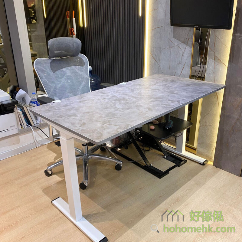 辦公室實拍全景，Morton電動升降枱灰色石紋枱面配白色金屬升降腳，正面角度可見T型腳座結構，枱旁有灰色辦公網椅，後方有Asus大熒幕電視，背景為金色邊框裝飾牆，木地板