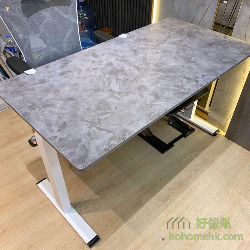 實拍照片，Morton電動升降枱灰色大理石仿石紋枱面俯視近距離拍攝，枱面四角有白色保護角，枱面下配白色金屬升降腳，後方可見辦公椅、辦公室背景及木地板