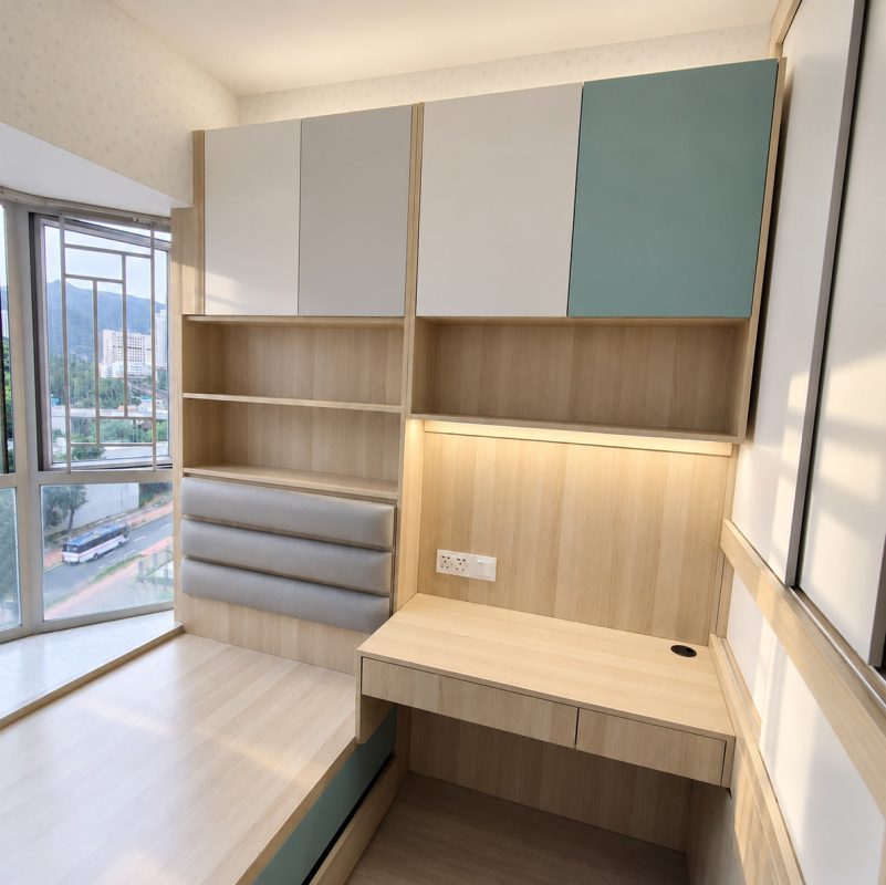 香港細單位訂造睡房全景，淺木色木紋為主色調，左側為地台床配薄荷綠抽屜面板，右側為一體式訂造組合包括書枱、開放書格、多個吊櫃（白色及薄荷綠門板）直至頂牆，書枱背板有LED燈帶及電源插座，旁邊大衣櫃趟門半開，窗外可見樓景及藍天，整體空間光線充足整齊。