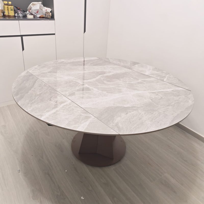 廚房旁的用餐位置放有一張完全打開的圓形岩板餐枱，亮光灰白石紋枱面分成多幅拼接，下方以啡灰色圓底中央座腳承托。
