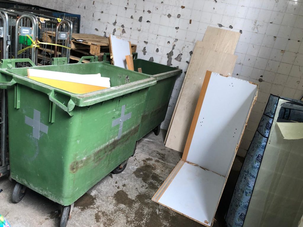 室內廢物存放區，綠色大型滾輪環保垃圾箱旁靠著多塊白色夾板及木色板材，牆壁磁磚剝落，傢俬廢料整齊靠牆放置