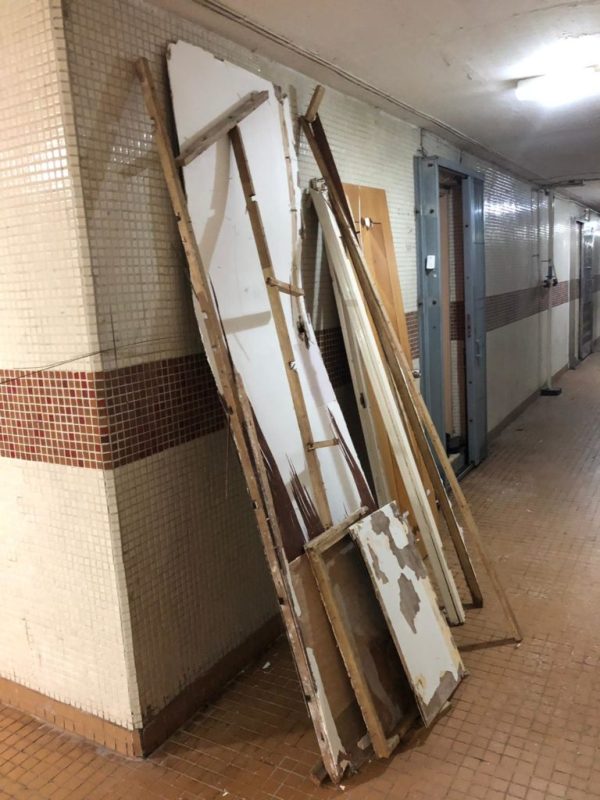 住宅大廈走廊磁磚牆邊，多塊拆除後的白色夾板及木框架靠牆豎放，走廊盡頭可見大廈門口及電燈，等待運載的清拆廢料