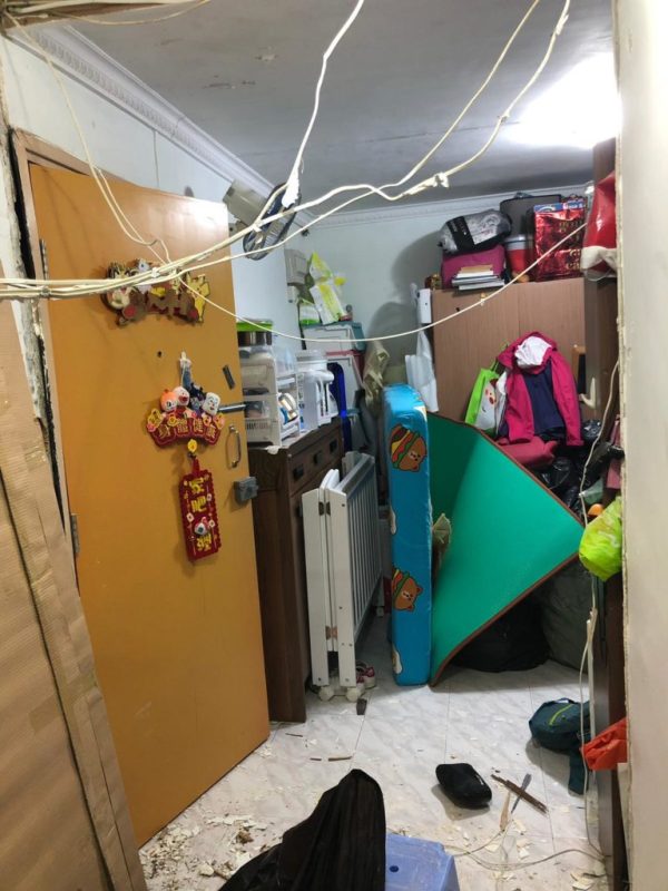 舊式住宅房間清拆中，牆壁局部拆除，天花橫跨白色電線，房內堆滿衣物袋、電暖爐、行李等舊物，地上散落碎料