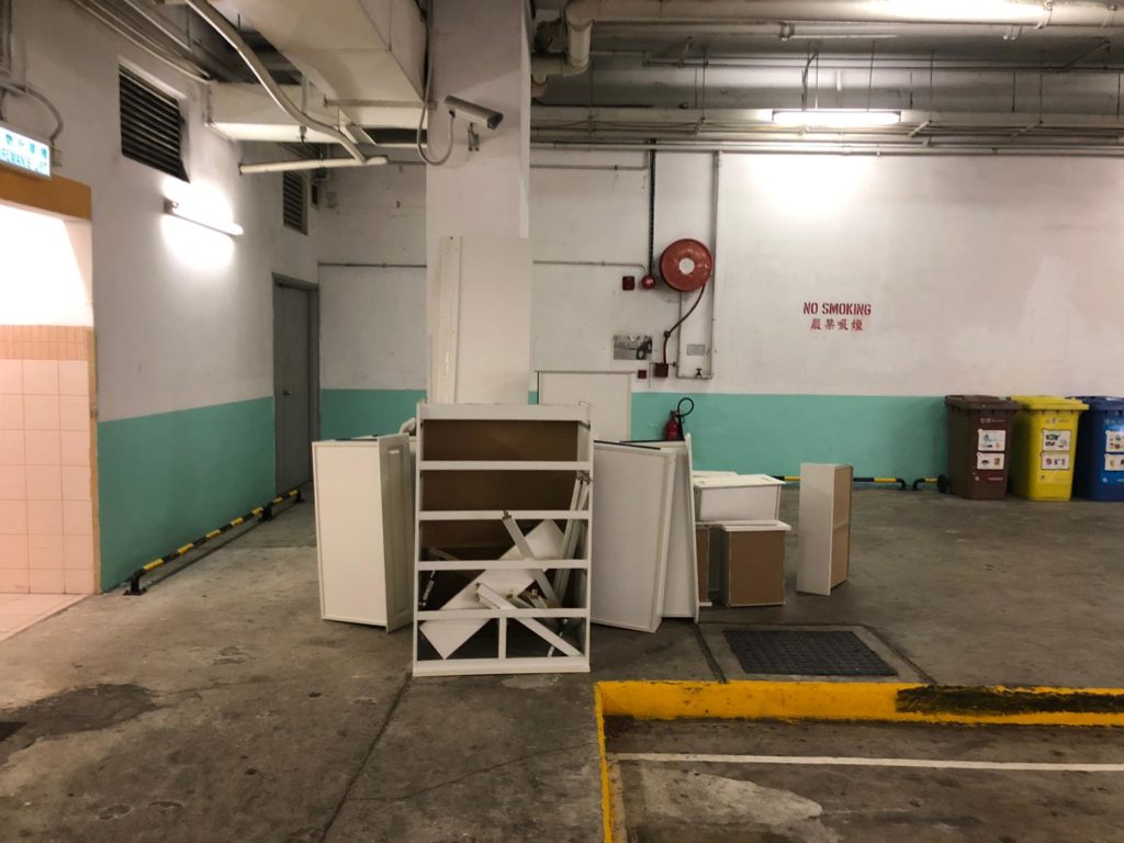 停車場內白色牆身前，多件白色系傢俬組件拆散後堆放地上，包括書架架身、抽屜側板及白色板材，場地有紅色消防裝置及英文禁煙告示