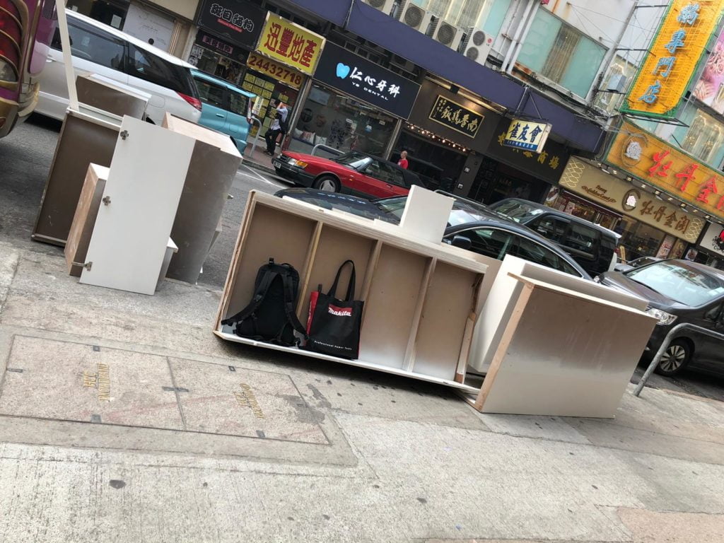 香港旺區街道行人路上，多件木色夾板傢俬組件橫放地上，背景為香港舊式商鋪招牌及街道車輛，師傅工具袋放於組件旁