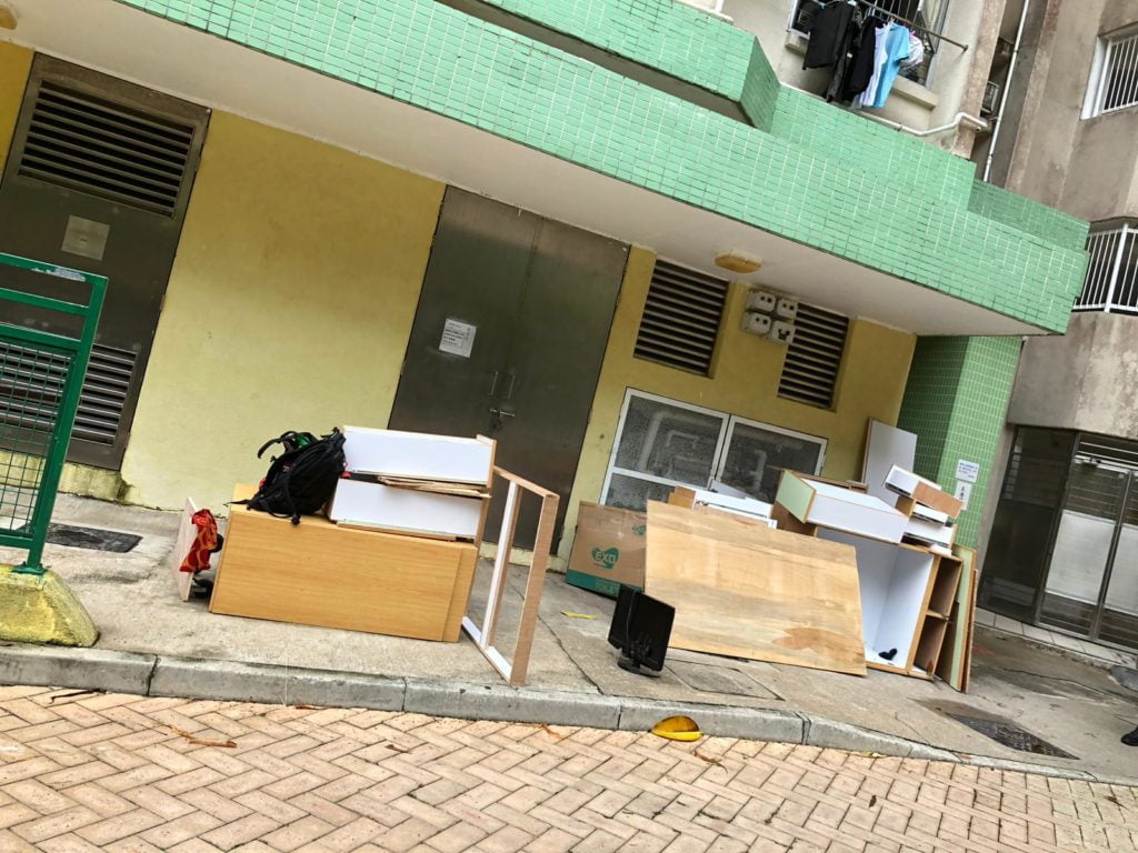 香港公共屋邨地下外牆旁，多件白色及木色夾板傢俬板材整齊疊放於行人路上，背景為黃綠色磁磚外牆及大廈出入口