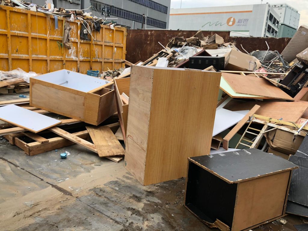 廢物收集站戶外場地，大量拆解後的傢俬板材、衣櫃組件及木材廢料堆放於大型黃色鐵箱旁，遠處可見商業大廈，天色陰沉