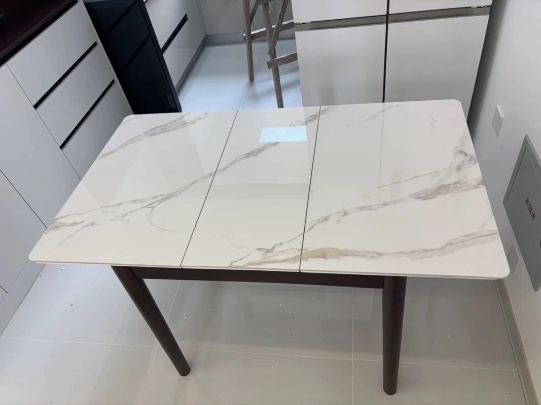 白色廚房內放有一張展開的岩板伸縮餐枱，三段亮面白色枱板帶灰啡雲石紋，圓角枱面下方配深啡色實木橫框及圓柱枱腳。