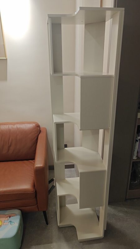 白色Peyton落地儲物書架側面實拍擺喺水泥地面門市展示區，5層階梯式錯位開放層板由頂到底清楚露出，左邊有壁燈射黃光，前面焦糖啡色皮梳化坐墊配黑色細腳，左下角有薄荷綠腳凳。