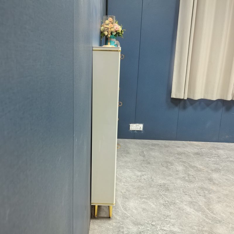 米白色超薄鞋櫃側面實拍，櫃身極纖薄近乎緊貼藍色布紋牆身，右邊為米色窗簾同白色雙位電源插座，櫃頂擺住金色燭台同粉紅小花瓶，底部為金色圓柱腳踩灰色雲石紋地板。