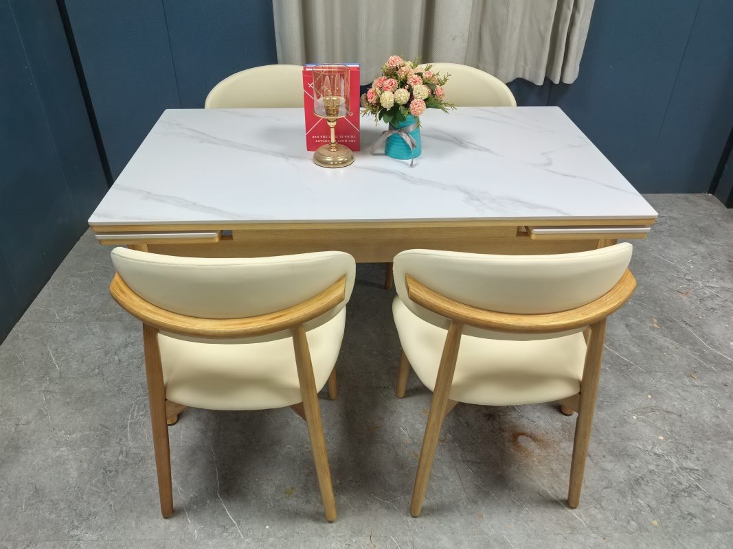 從後方高角度拍攝Reese白色岩板伸縮餐枱，前景並排兩張米白色軟墊弧背餐椅，原木色圓弧扶手、木腳與枱架一致。