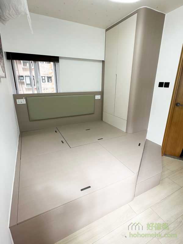 主人房的設計重點在於因應套廁門口的位置，特別訂造了斜角地台，避免出入廁所時的不便。此外，卡其色的地台床與圓邊衣櫃相互呼應，不僅提升了安全性，還增添了居室的溫馨感。床頭綠色軟墊的設計，更為整體空間帶來了一抹柔和的色彩。