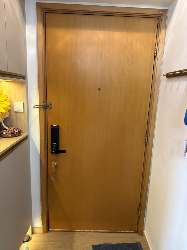 翻新後米黃木門入戶全景、門框回復木色、旁邊有淺灰廚櫃連檯面擺住向日葵擺設、右邊白牆掛住金色四鉤木製衣帽架、門上裝有黑色 Samsung 智能鎖連金色流蘇、地下有淺色地板。