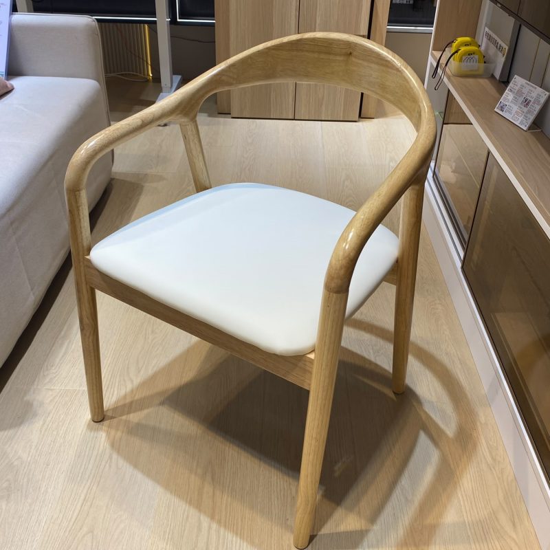 門市木地板上近距離拍攝原木色舒美特餐椅，圓滑弧背連接兩邊扶手及前腳，寬身米白皮坐墊置於木框內，背景為淺木色門市櫃。